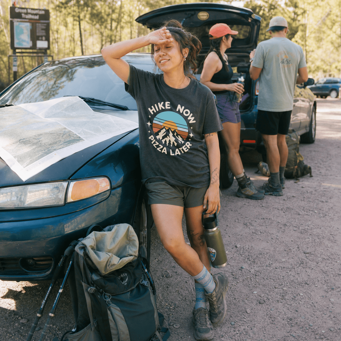 Woman resting against a car smiles while wearing a "Hike Now Pizza Later Shirt" featuring mountains and a sunset design.