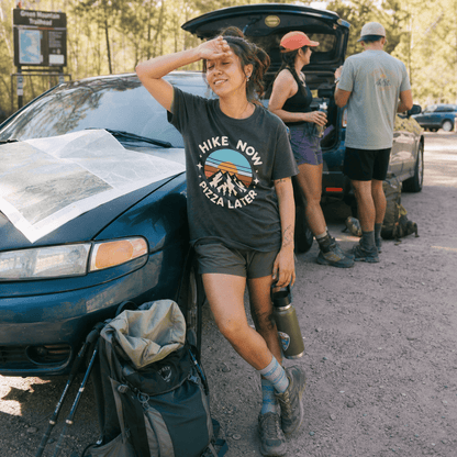 Woman resting against a car smiles while wearing a "Hike Now Pizza Later Shirt" featuring mountains and a sunset design.