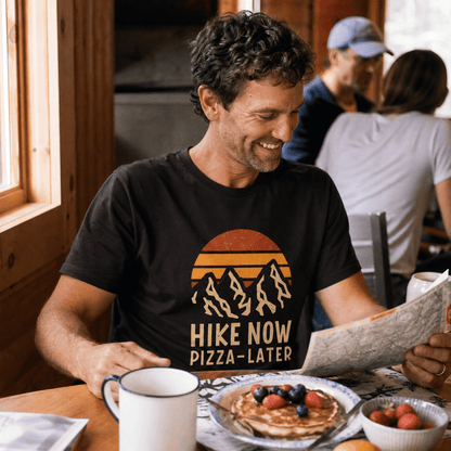 Man seated at a table, smiling while looking at a map, wearing a "Hike Now Pizza Later Shirt" with a mountain and sunset grap