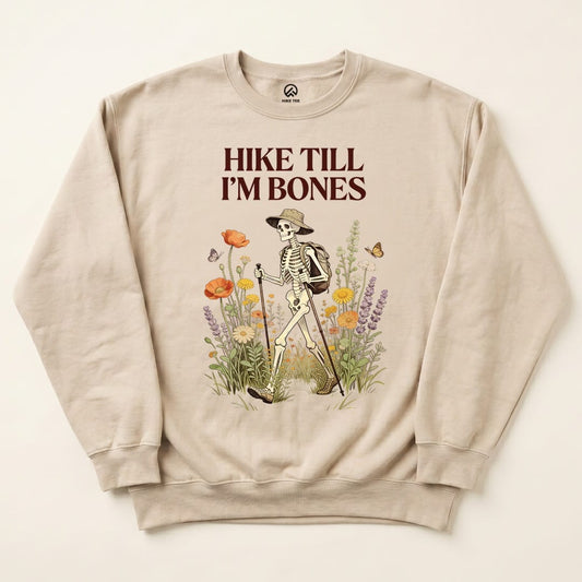 A graphic sweatshirt featuring a skeleton hiking with a backpack and the slogan "Hike Till I'm Bones" surrounded by flowers and butterflies.