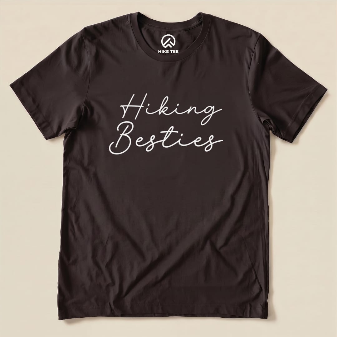 A graphic t-shirt in dark color featuring the phrase "Hiking Besties" in a fun, cursive font displayed prominently on the front.