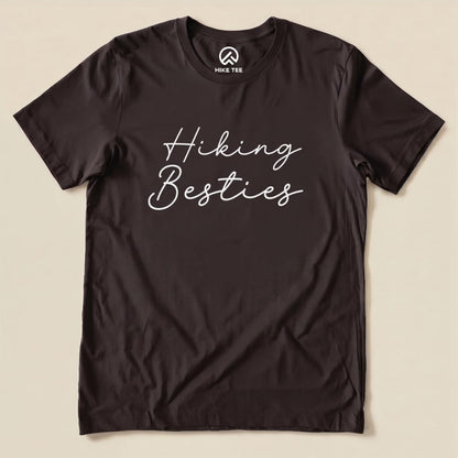 A graphic t-shirt in dark color featuring the phrase "Hiking Besties" in a fun, cursive font displayed prominently on the front.
