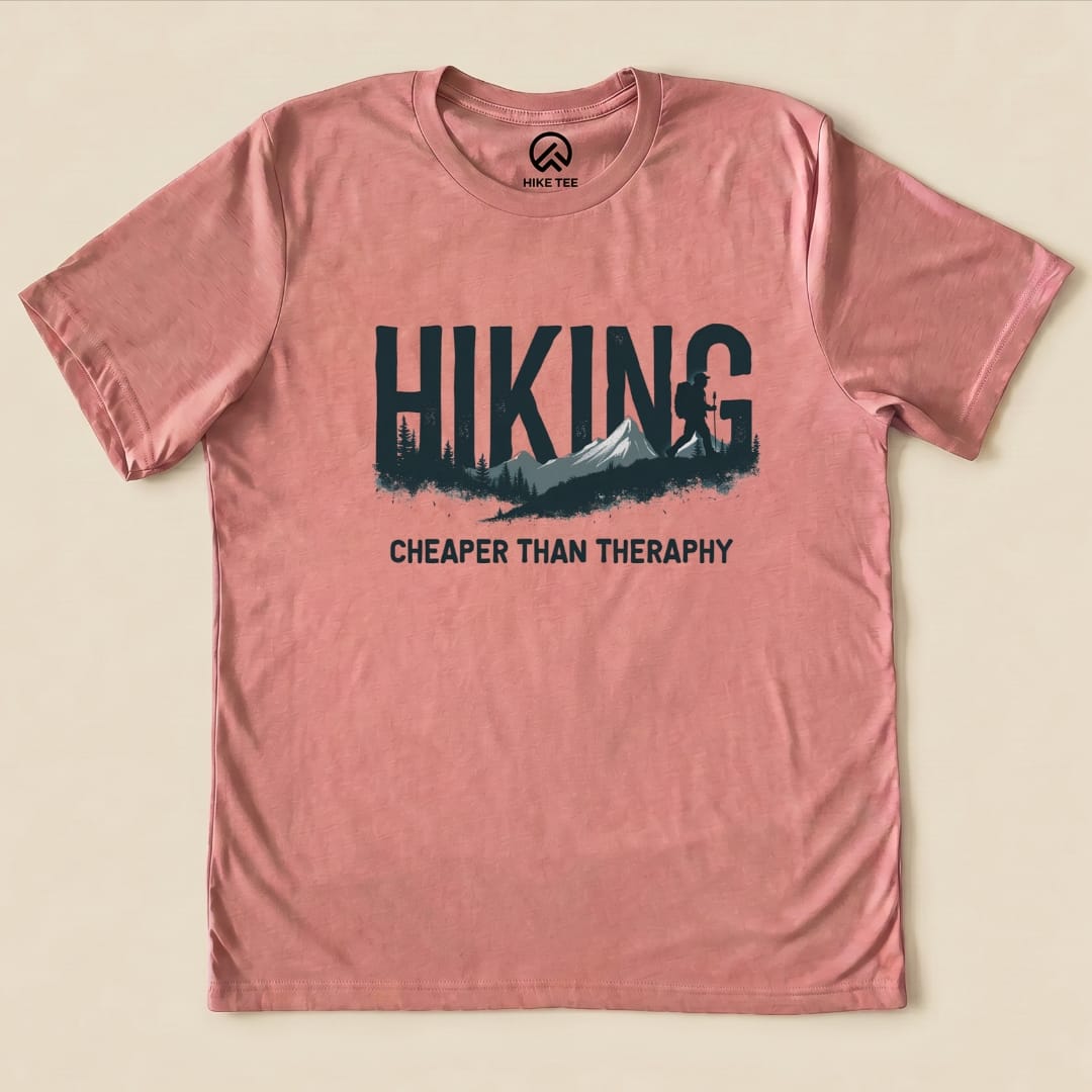 A pink graphic t-shirt featuring the slogan "Hiking - Cheaper than Therapy" with a mountain and hiker design.
