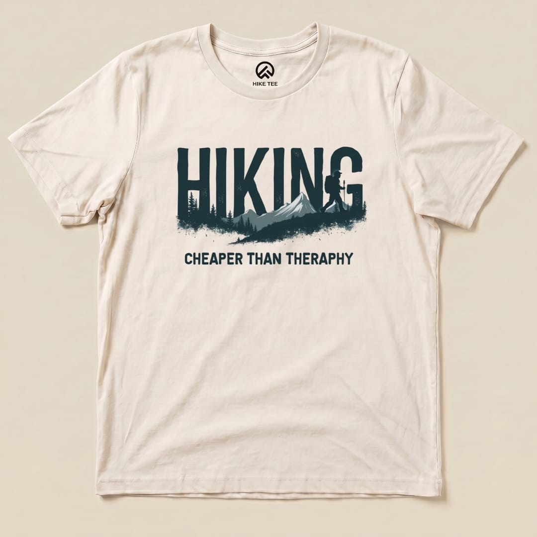 A cream-colored graphic t-shirt featuring the phrase "Hiking - Cheaper than Therapy" alongside a mountain and forest design.