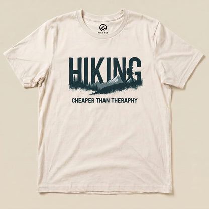 A cream-colored graphic t-shirt featuring the phrase "Hiking - Cheaper than Therapy" alongside a mountain and forest design.
