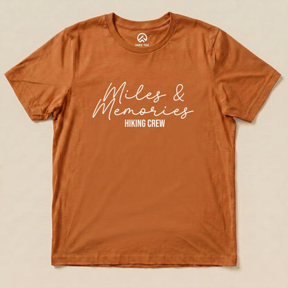 A flat view of an orange graphic t-shirt featuring the words "Miles & Memories" with "Hiking Crew" underneath.