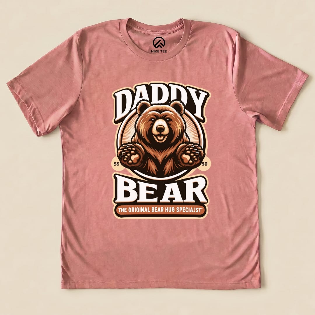 A graphic t-shirt in light pink featuring a playful bear design with the text "Hug Daddy Bear Shirt" and the slogan "THE ORIGINAL BEAR HUG SPECIALIST.