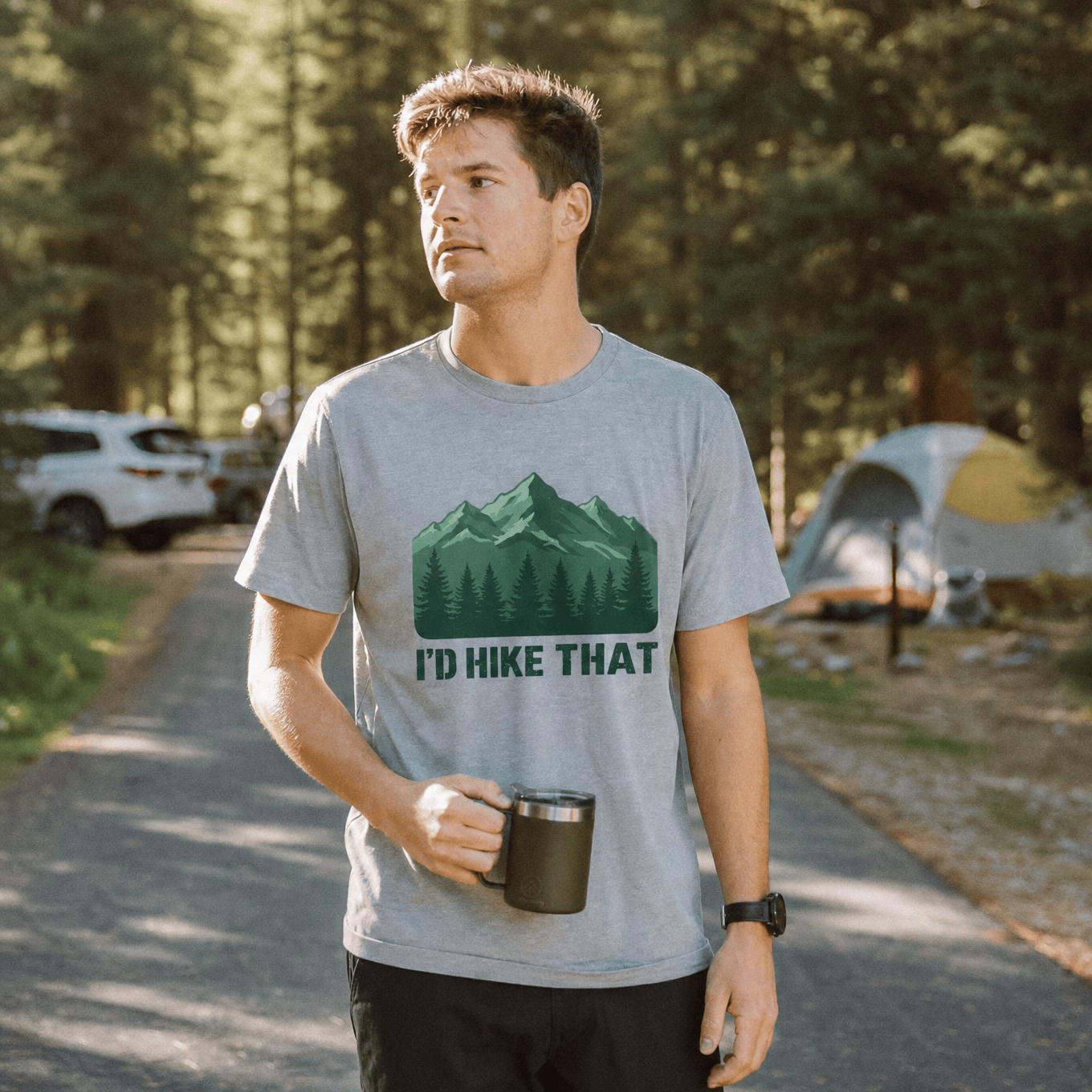 A man stands on a forest path wearing a gray graphic t-shirt featuring the text "I'D HIKE THAT" above a mountain and tree design.