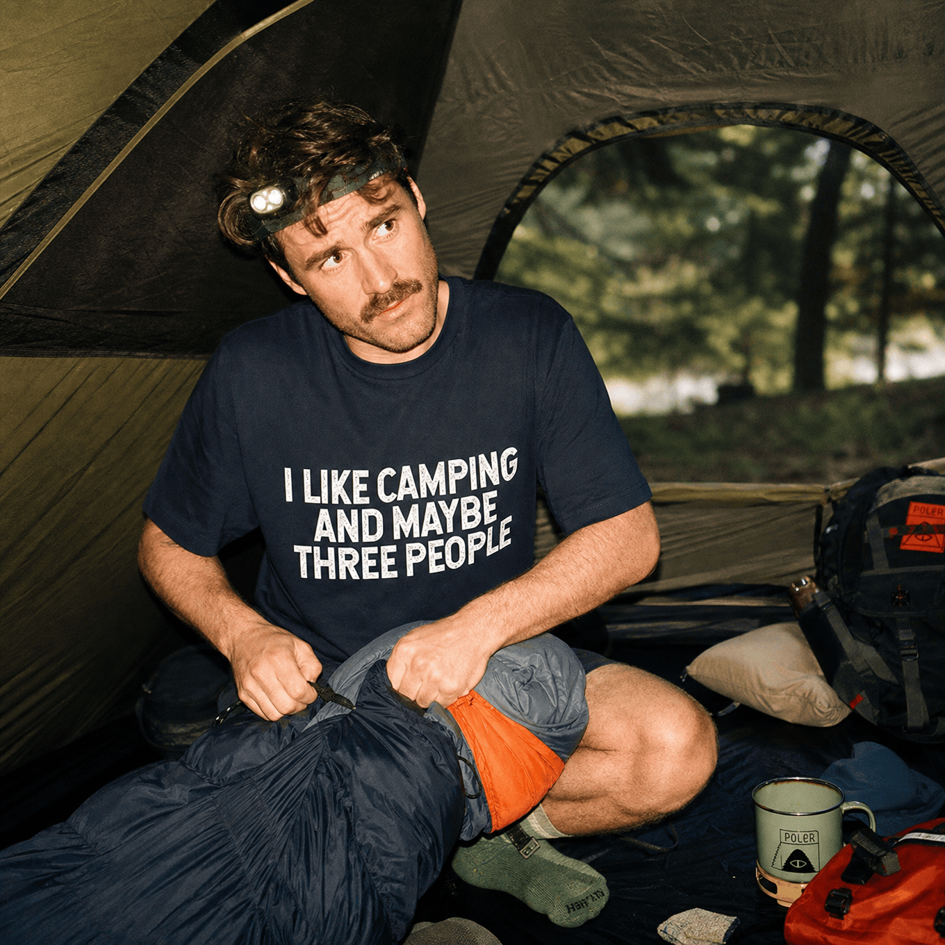 A man sits inside a tent, wearing a graphic t-shirt that reads, "I LIKE CAMPING AND MAYBE THREE PEOPLE," as he packs a sleeping bag.