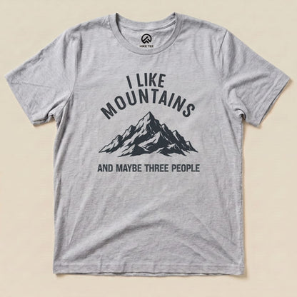 A gray graphic t-shirt featuring the text "I Like Mountains" above a mountain graphic, with the phrase "AND MAYBE THREE PEOPLE" below.