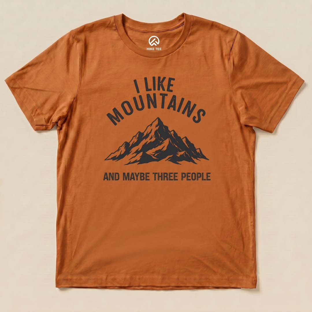 A flat view of the "I Like Mountains Shirt" featuring a graphic of a mountain range with the text "I LIKE MOUNTAINS AND MAYBE THREE PEOPLE" on an orange background.