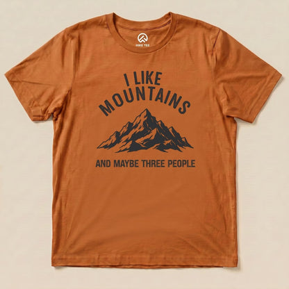 A flat view of the "I Like Mountains Shirt" featuring a graphic of a mountain range with the text "I LIKE MOUNTAINS AND MAYBE THREE PEOPLE" on an orange background.