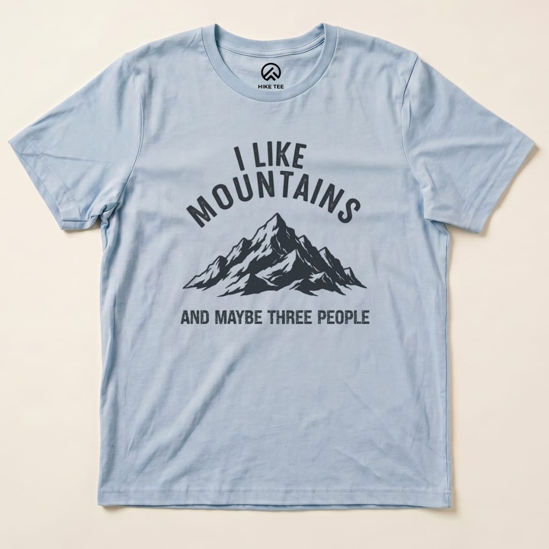 A light blue graphic t-shirt featuring the phrase "I Like Mountains" above a mountain illustration, with "AND MAYBE THREE PEOPLE" below.