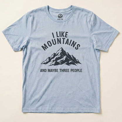 A light blue graphic t-shirt featuring the phrase "I Like Mountains" above a mountain illustration, with "AND MAYBE THREE PEOPLE" below.