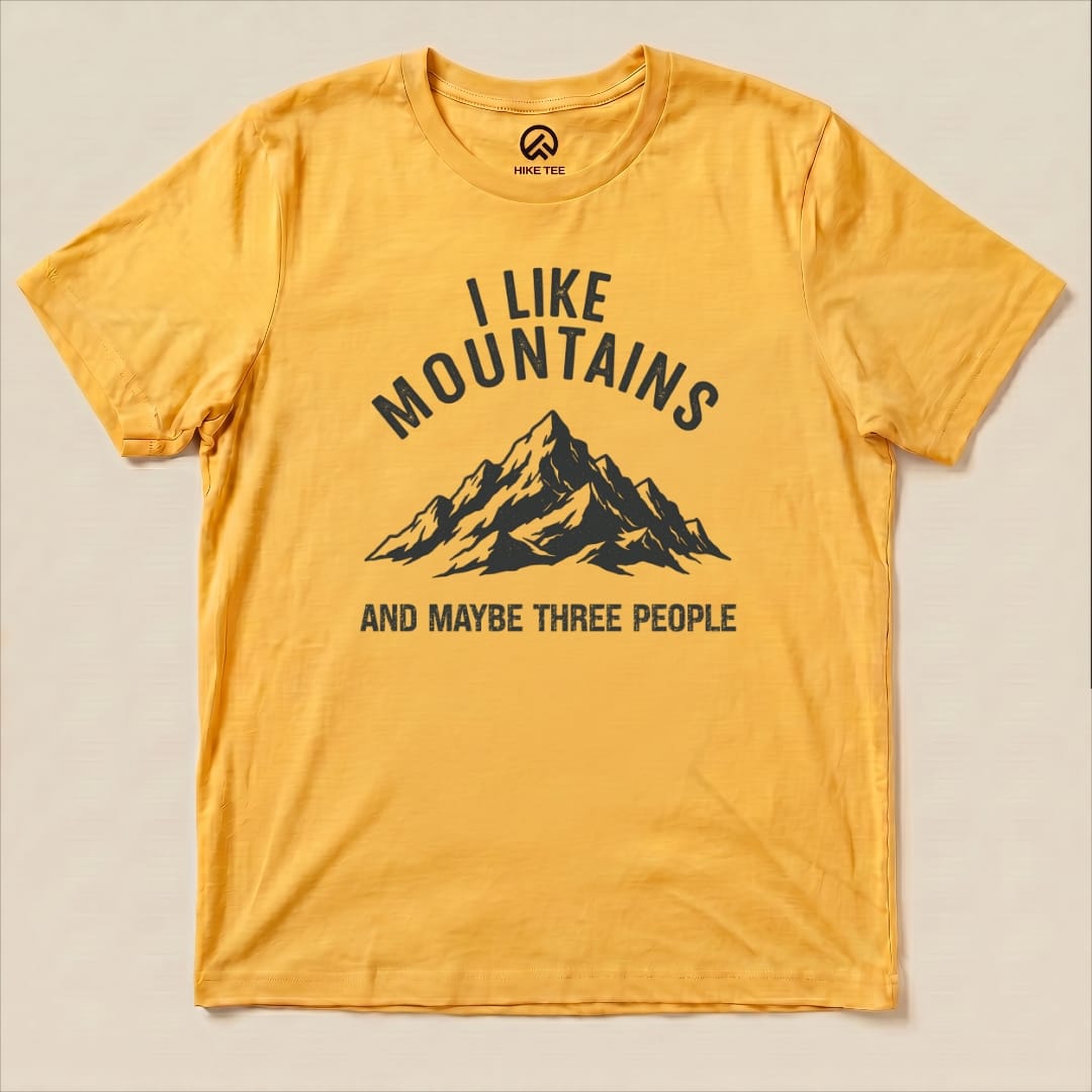 A graphic t-shirt featuring a mountain illustration with the text "I Like Mountains AND MAYBE THREE PEOPLE" in a bold design.