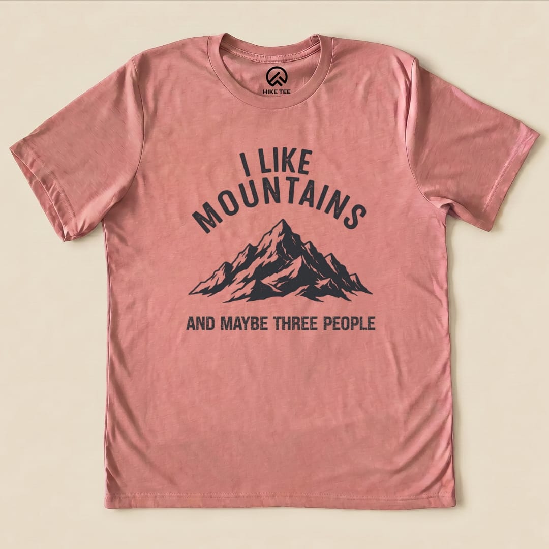 A graphic t-shirt in a soft pink color featuring the phrase "I Like Mountains" above a mountain graphic and "AND MAYBE THREE PEOPLE.
