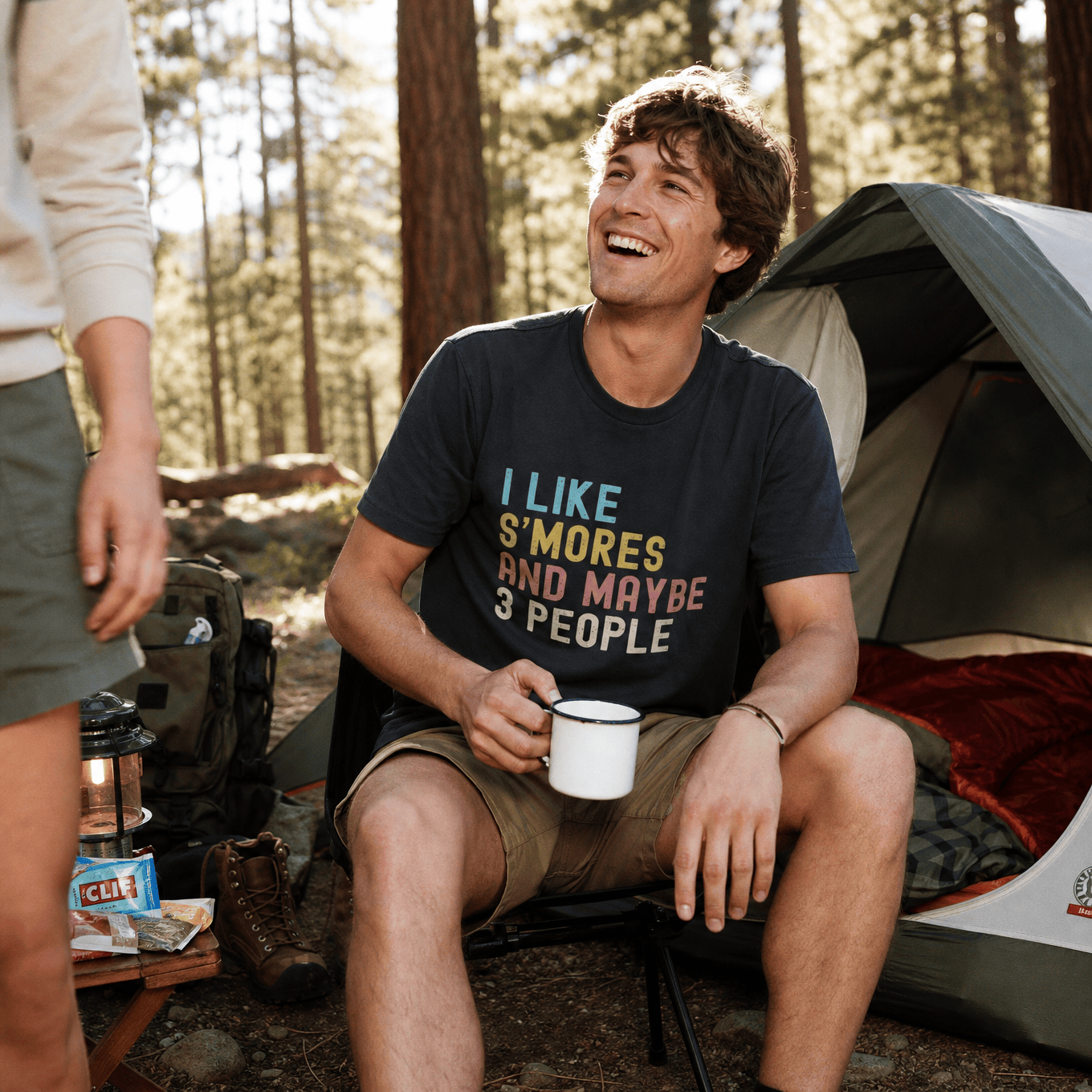 A man sitting by a tent in the woods smiles while wearing a graphic t-shirt that says "I like S'mores and maybe 3 people.