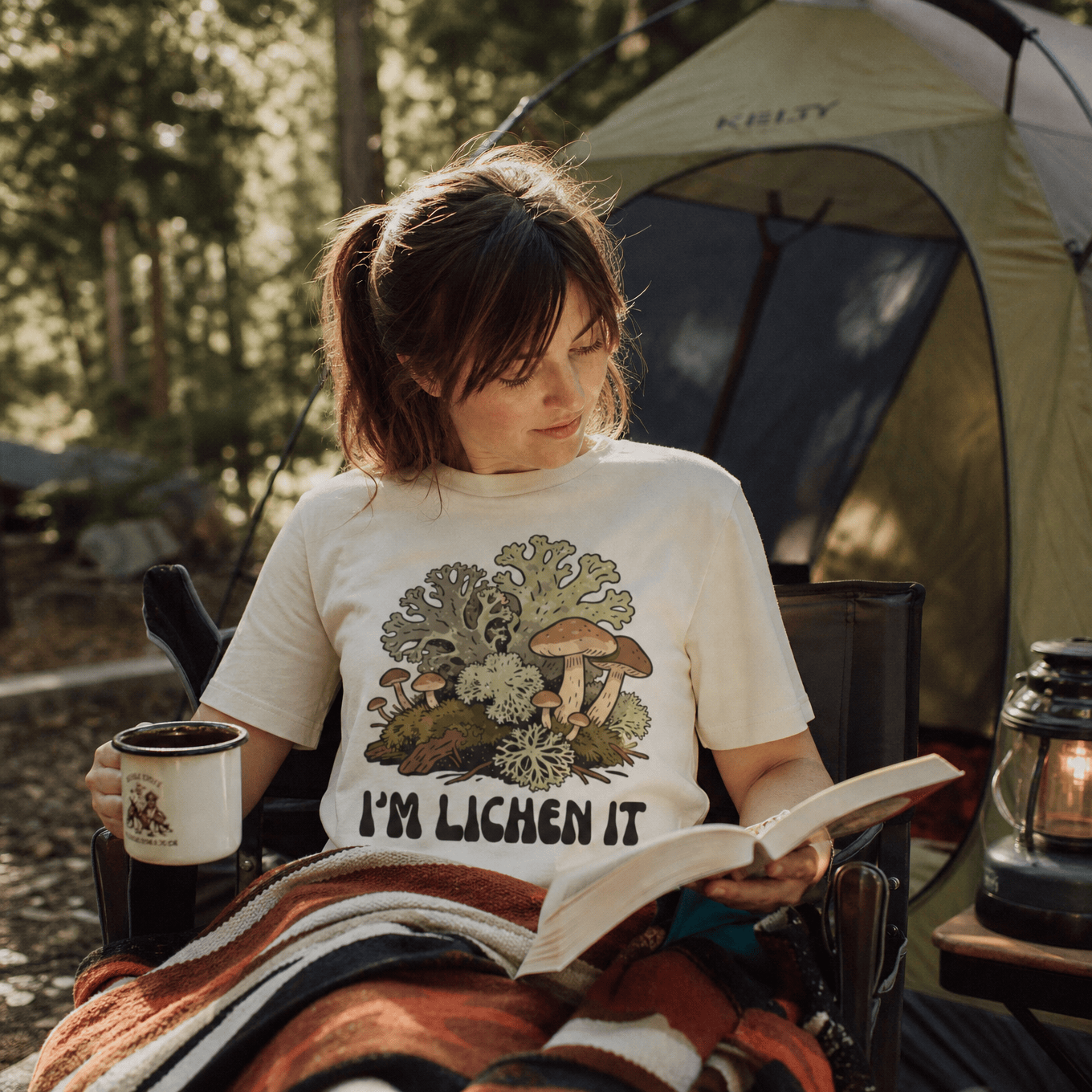 A woman sits in a camping chair, reading a book while wearing a graphic t-shirt featuring the phrase "I'm Lichen It" and whimsical mushroom designs.