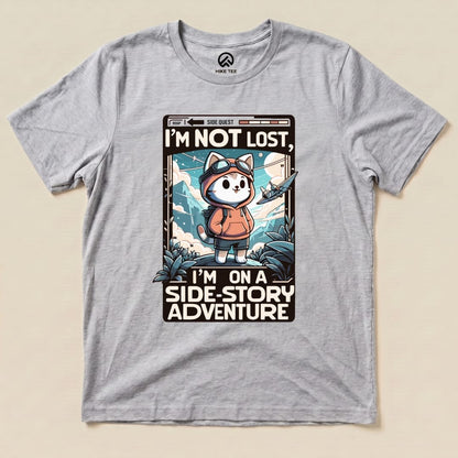 Gray graphic t-shirt featuring a cartoon cat in a hoodie with goggles, stating "I'm Not Lost, I'm on a Side-Story Adventure.