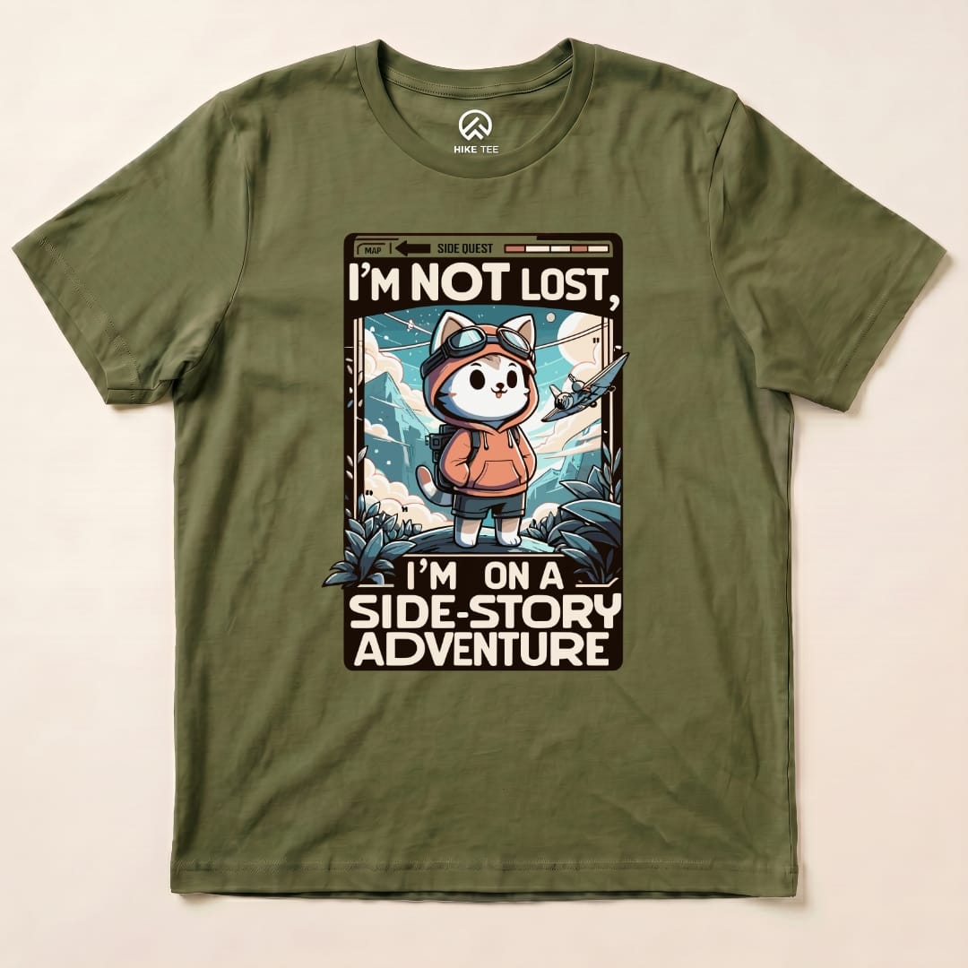 A graphic t-shirt in olive green featuring a cartoon cat in a hoodie, with the phrase "I'M NOT LOST, I'M ON A SIDE-STORY ADVENTURE.