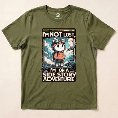 A graphic t-shirt in olive green featuring a cartoon cat in a hoodie, with the phrase "I'M NOT LOST, I'M ON A SIDE-STORY ADVENTURE.