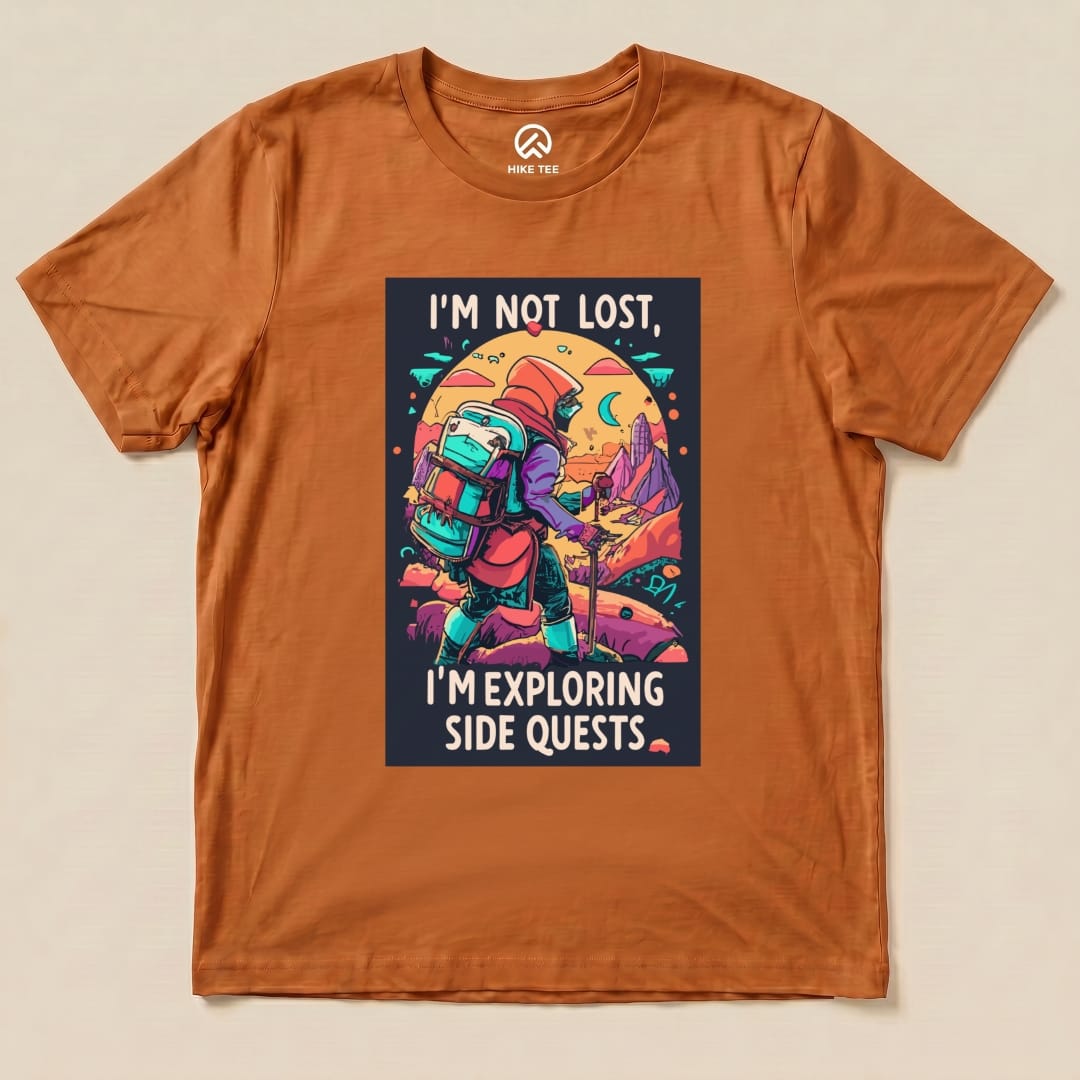 A graphic t-shirt featuring an illustration of a hiker with a backpack and the text "I'm Not Lost, I'm Exploring Side Quests" on an orange background.