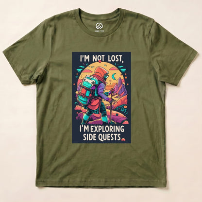 A graphic t-shirt in olive green featuring the phrase "I'm Not Lost, I'm Exploring Side Quests" with an illustration of a hiker.