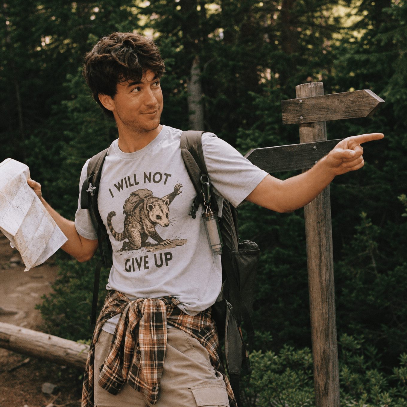 A man stands in a forest, holding a map while wearing a graphic t-shirt featuring an opossum and the slogan "I WILL NOT GIVE UP".