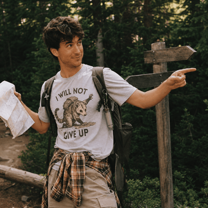 A man stands in a forest, holding a map while wearing a graphic t-shirt featuring an opossum and the slogan "I WILL NOT GIVE UP".