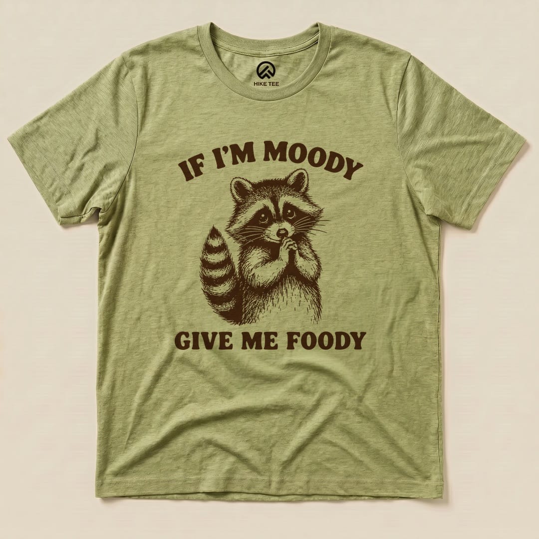 The graphic t-shirt displays a raccoon with the text "IF I'M MOODY GIVE ME FOODY" printed in a playful design on an olive green background.