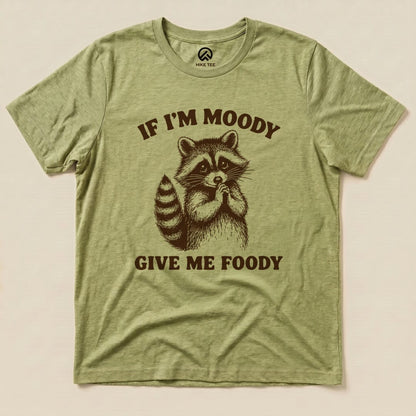 The graphic t-shirt displays a raccoon with the text "IF I'M MOODY GIVE ME FOODY" printed in a playful design on an olive green background.