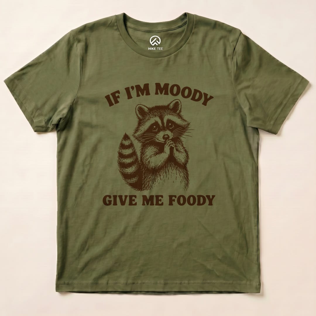 A graphic t-shirt in olive green featuring a raccoon and the text "IF I'M MOODY GIVE ME FOODY" in bold letters.