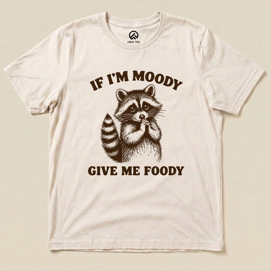 A graphic t-shirt displaying a cute raccoon with the text "IF I'M MOODY GIVE ME FOODY" printed in bold.