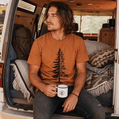 A man sits in the back of a van, wearing an "In The Forest Shirt" featuring a tree graphic and the phrase about finding one's soul.