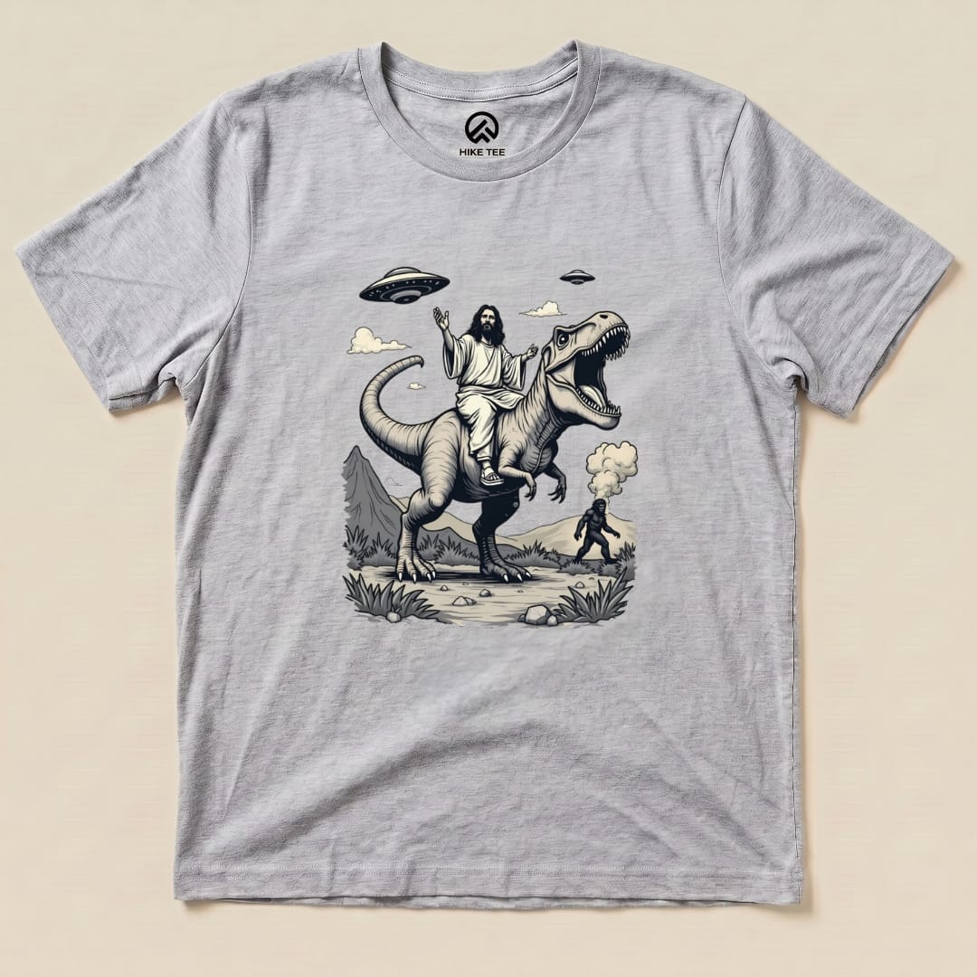 A graphic t-shirt featuring Jesus riding a T-rex, with Bigfoot in the distance, surrounded by mountains and UFOs.