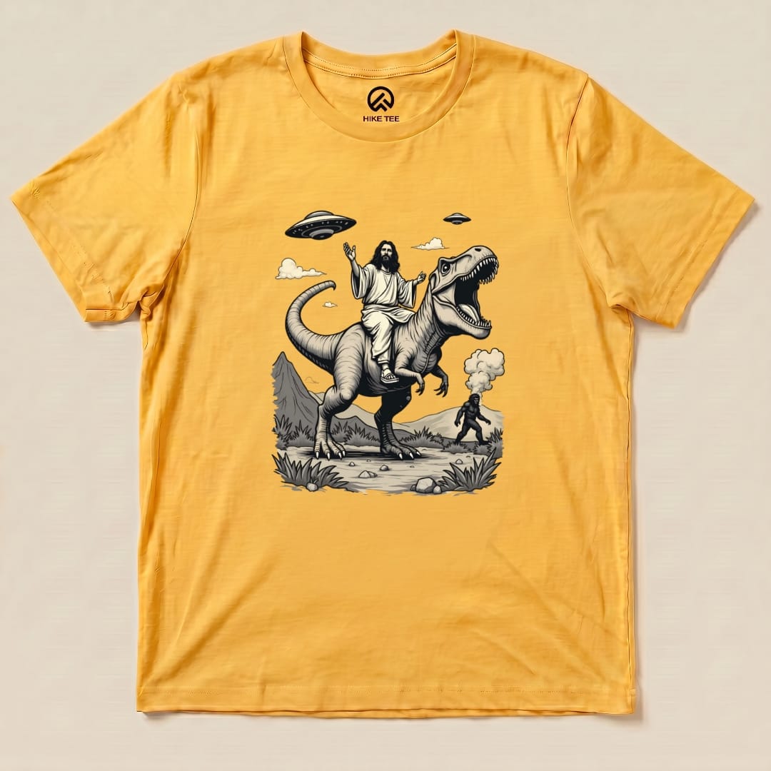 A graphic t-shirt in yellow featuring Jesus riding a T-rex, with Bigfoot and UFOs in the background.