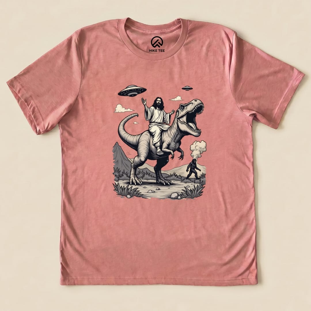 This graphic t-shirt features an illustration of Jesus riding a T-rex with Bigfoot in the background, set against a whimsical landscape.