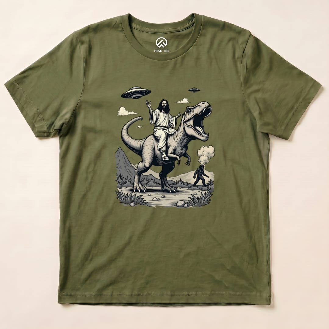 A graphic t-shirt featuring Jesus riding a T-rex, with Bigfoot in the background, and UFOs above in a whimsical design.
