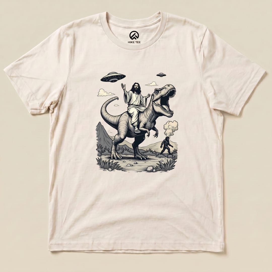 A graphic t-shirt featuring Jesus riding a T-rex with Bigfoot in the background and UFOs in the sky, titled "Jesus with T-rex and Bigfoot Shirt.