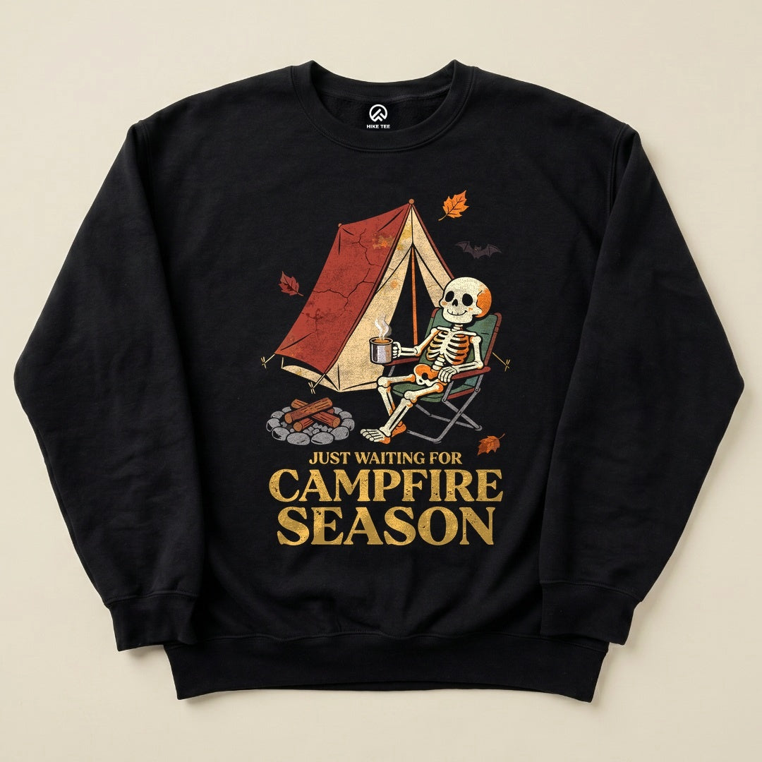 A black sweatshirt featuring a humorous graphic of a skeleton sitting by a tent, holding a drink, and the text "JUST WAITING FOR CAMPFIRE SEASON.