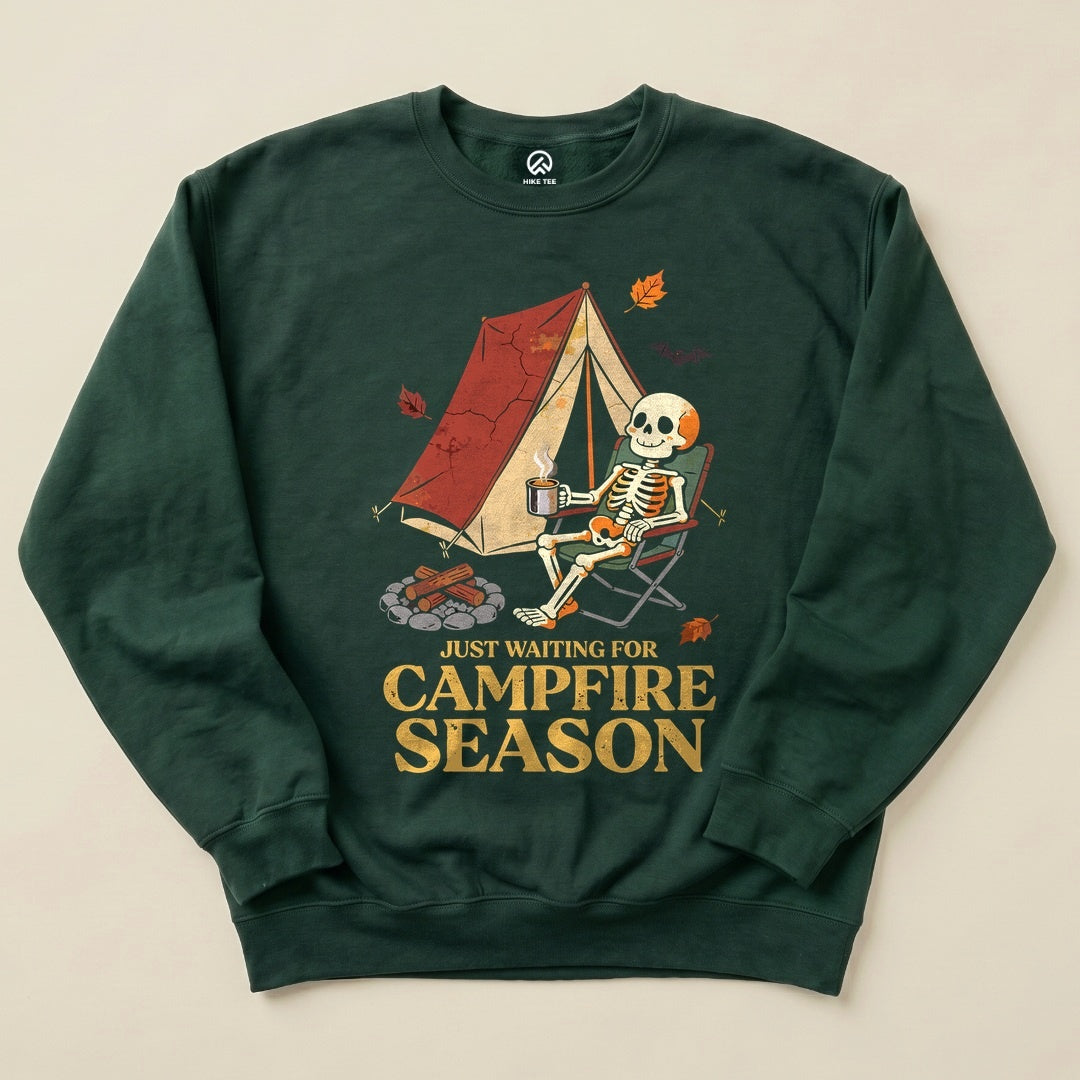A graphic sweatshirt featuring a skeleton lounging in a camping chair next to a tent and fire, with "Just Waiting for Campfire Season" printed below.