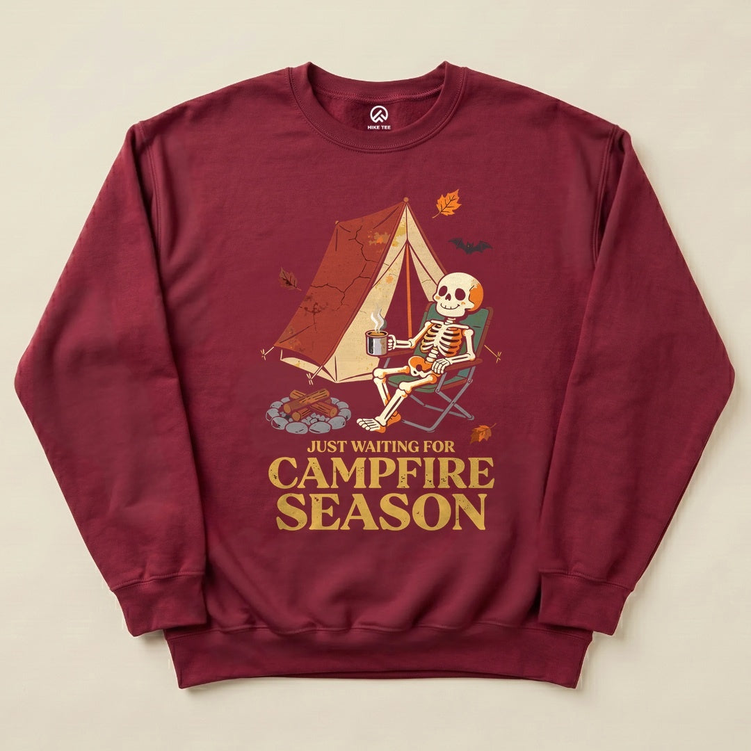 A maroon graphic sweatshirt features a skeleton relaxing in a camping chair next to a tent, with the text "Just Waiting for Campfire Season".