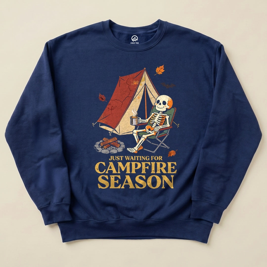 A navy sweatshirt displays a graphic of a skeleton sitting in a chair by a tent, with the words "Just Waiting for Campfire Season.