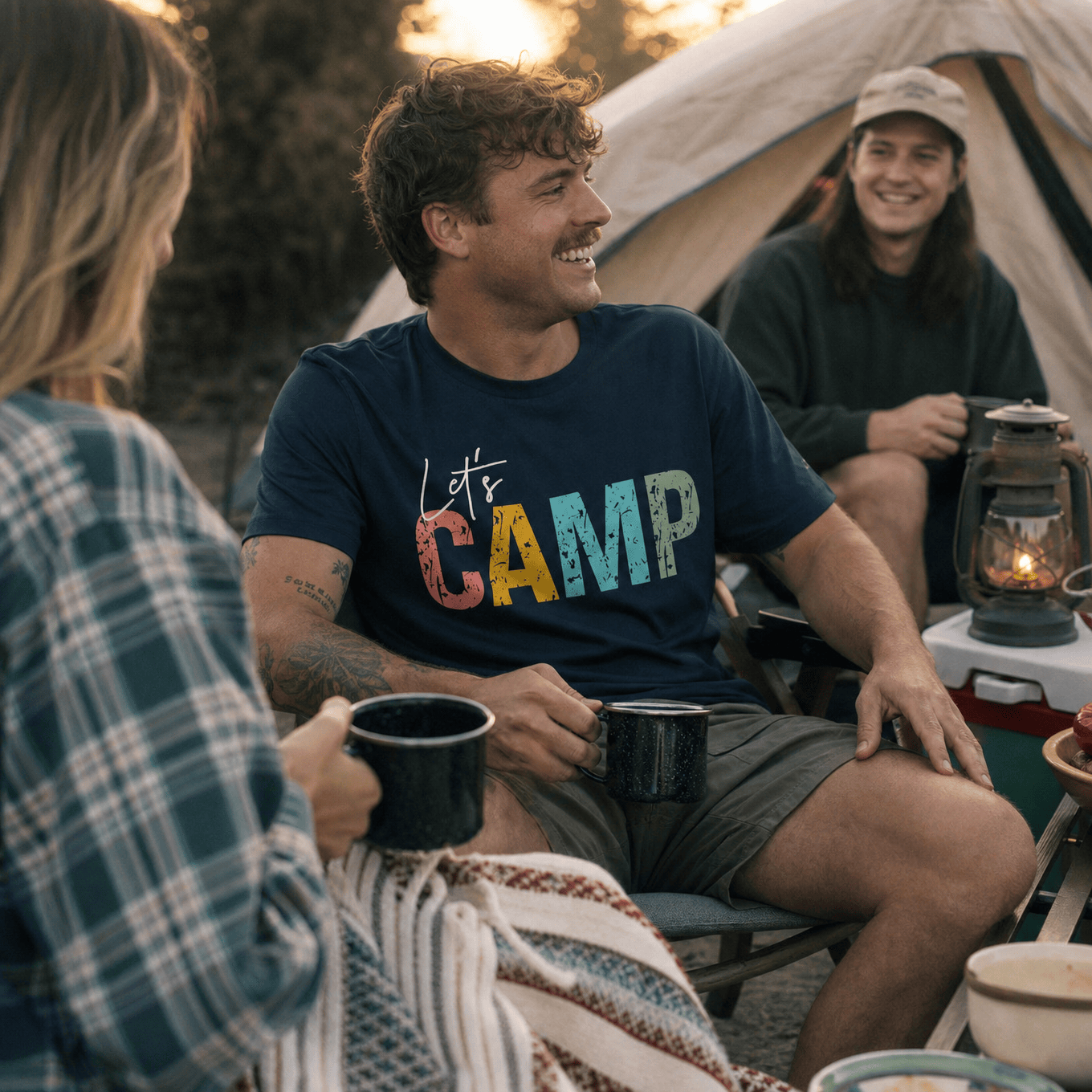 A man smiles while sitting by a campfire, wearing a navy graphic t-shirt that says "Let's Camp" in colorful letters.