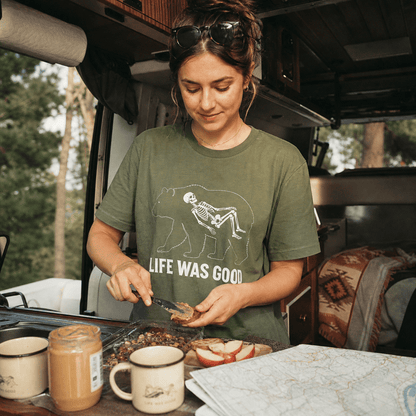A woman prepares food in a van wearing a green graphic t-shirt featuring a bear and a skeleton with the words "Life Was Good.