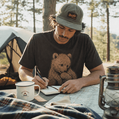 A man sitting at a camping table writes in a notebook while wearing the Little Bear Shirt featuring a printed graphic of a cute bear.