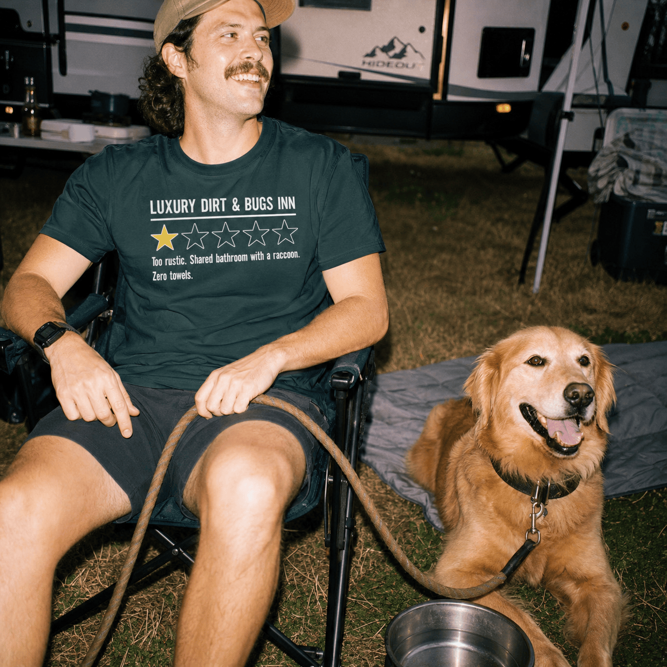 A man sits in a camping chair with a golden retriever, wearing a graphic t-shirt featuring the slogan "Luxury Dirt & Bugs Inn" and a humorous review.