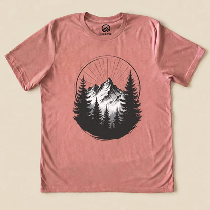 The Monochrome Mountains Shirt features a striking graphic of mountains and pine trees within a circular design on a pink background.