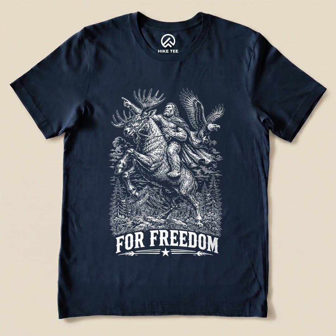 A navy graphic t-shirt features a comical design of Bigfoot riding a moose beneath the slogan "FOR FREEDOM".