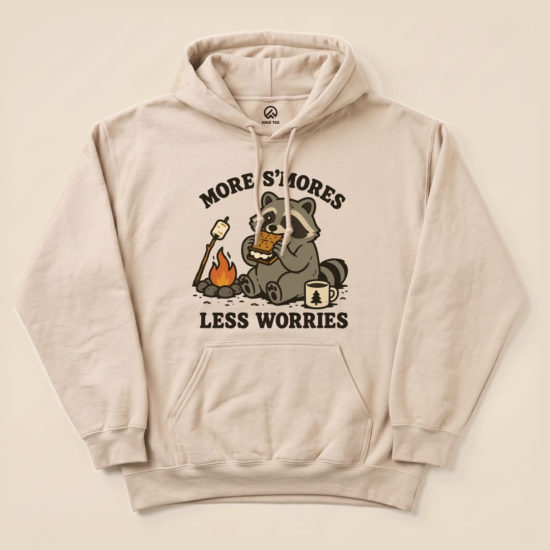A beige hoodie features a playful graphic of a raccoon enjoying s'mores by a campfire with the text "More S'mores Less Worries.
